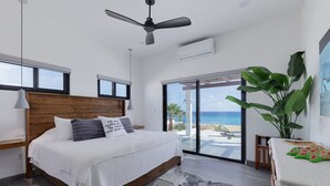 A modern bedroom with a large bed, a ceiling fan, and a view of the ocean through a sliding glass door.