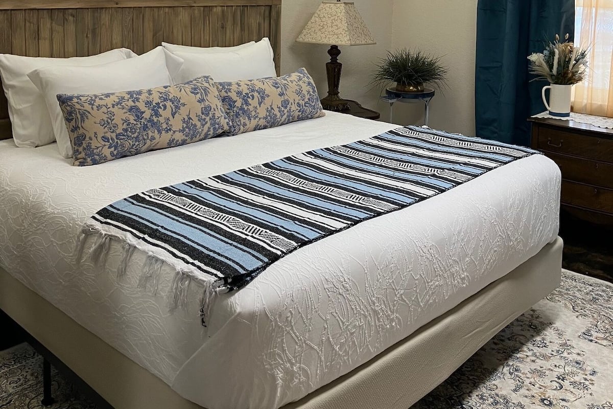 Uma cama bem arrumada com colcha estampada, cabeceira e mesa de cabeceira com luminária e planta.