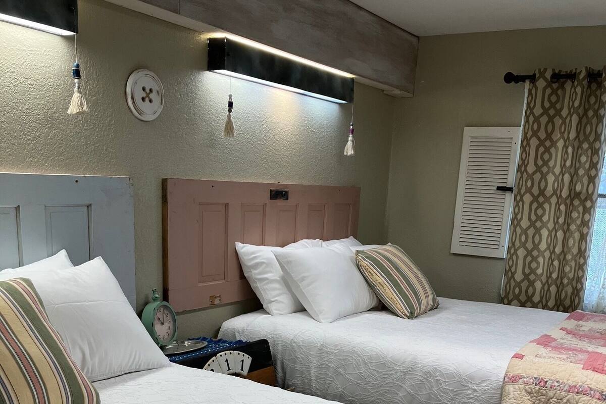Quarto de hotel com duas camas, cada uma com colcha e travesseiros brancos, uma cabeceira de madeira, um candeeiro fixado na parede e uma janela com cortinas estampadas.