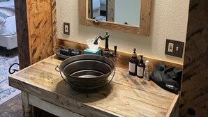 Une salle de bain avec un meuble-lavabo en bois, un grand miroir et un lavabo avec une cuve. On y trouve des articles de toilette sur le comptoir et une poubelle en dessous.
