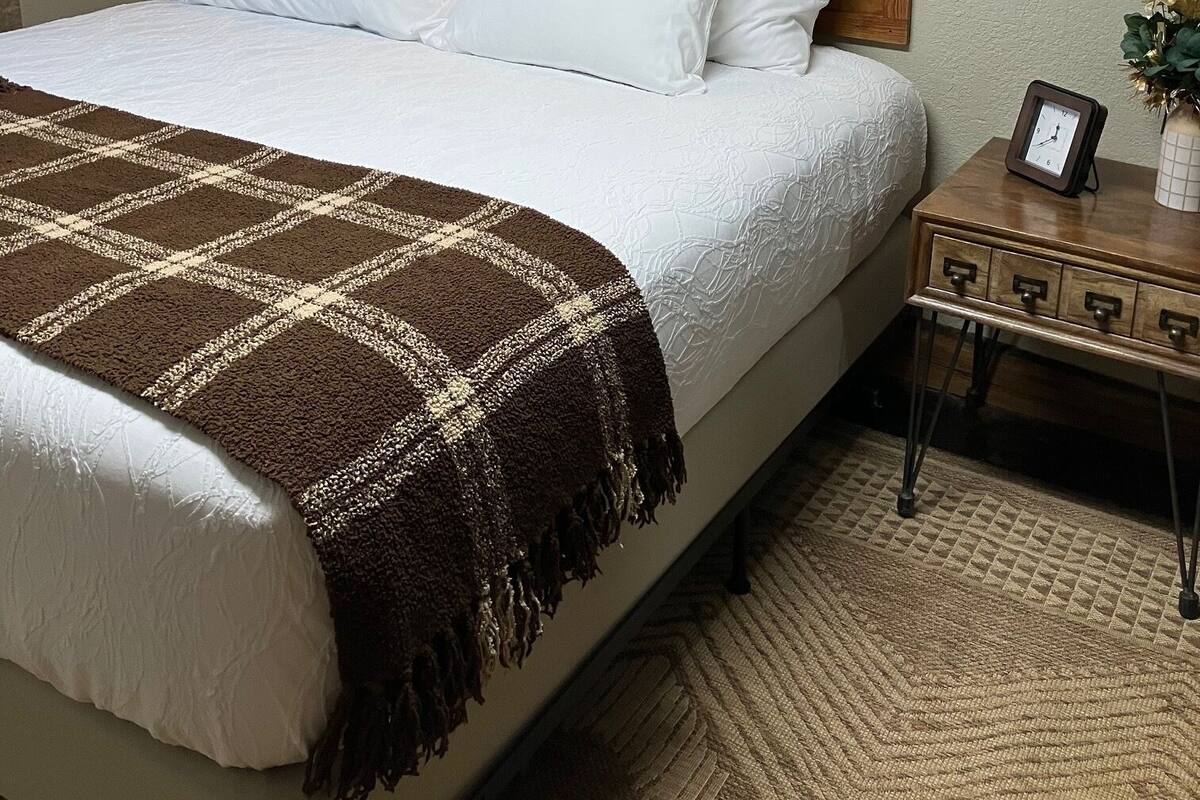 Cama bem arrumada com manta xadrez marrom e branca, cabeceira de madeira e mesa de cabeceira com relógio e um vaso de flores.