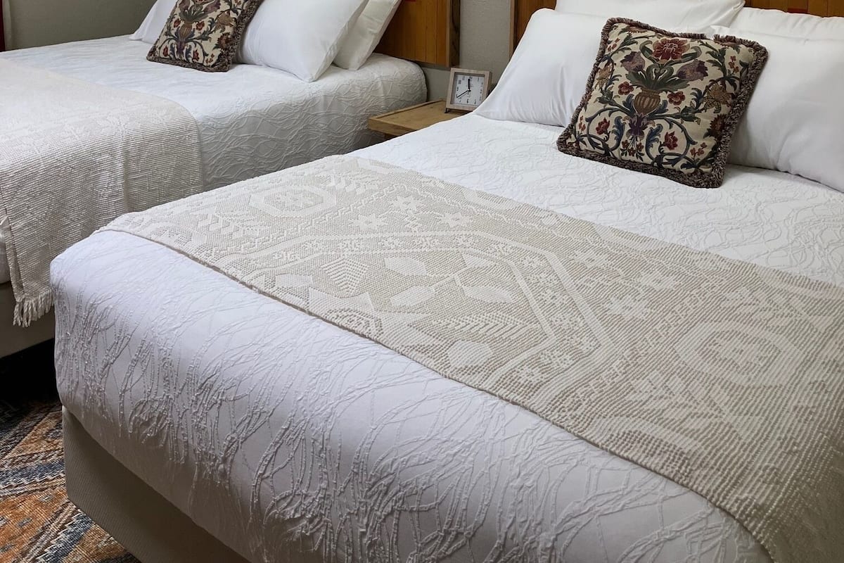 Quarto de hotel com duas camas, cada uma com roupa de cama branca e almofadas decorativas. Há luminárias fixadas na parede e um quadro emoldurado.