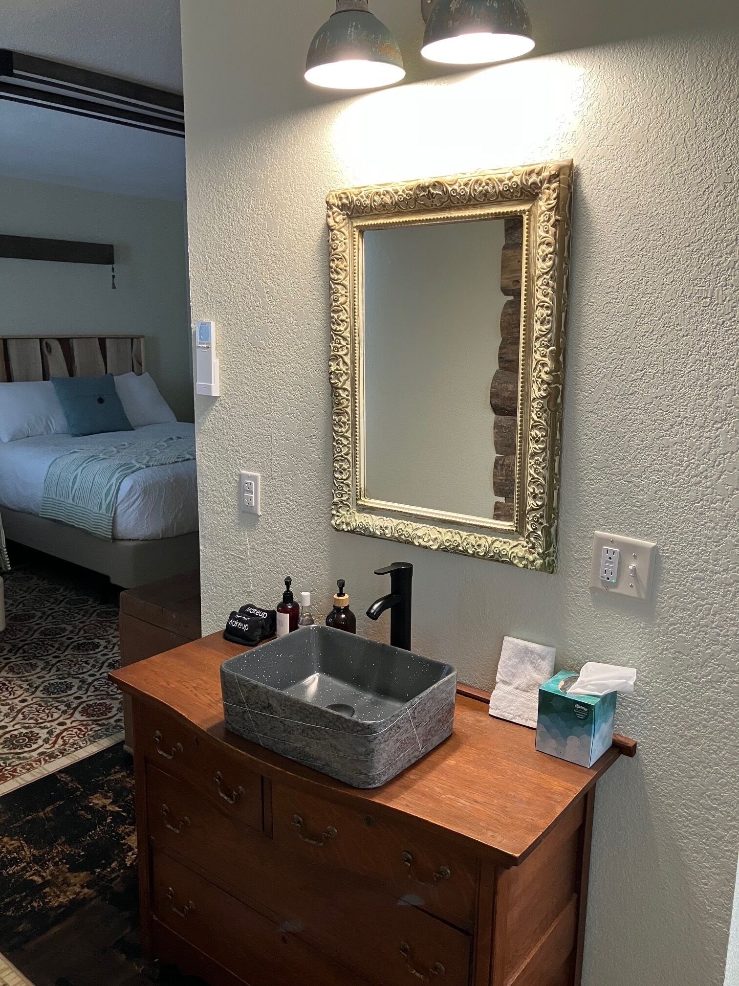 A bathroom with a marble sink, a wooden vanity, a mirror with an ornate frame, and a bed in the adjacent room.