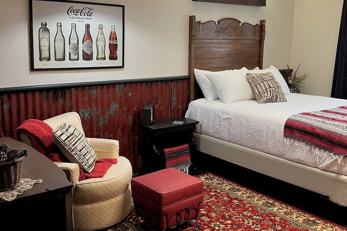Quarto de hotel com cama, poltrona, tapete vermelho, luminária fixada na parede e um anúncio emoldurado da Coca-Cola.