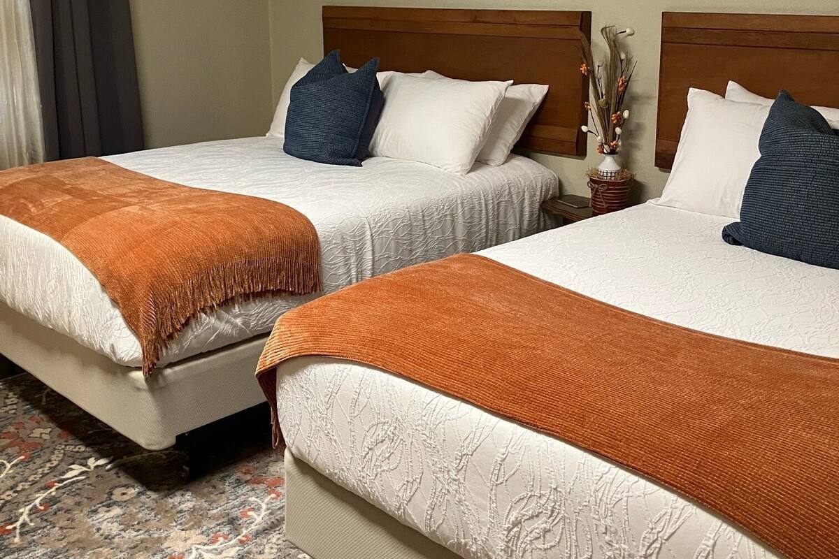 Quarto de hotel com duas camas, cada uma com cabeceira de madeira, roupa de cama branca e manta laranja.