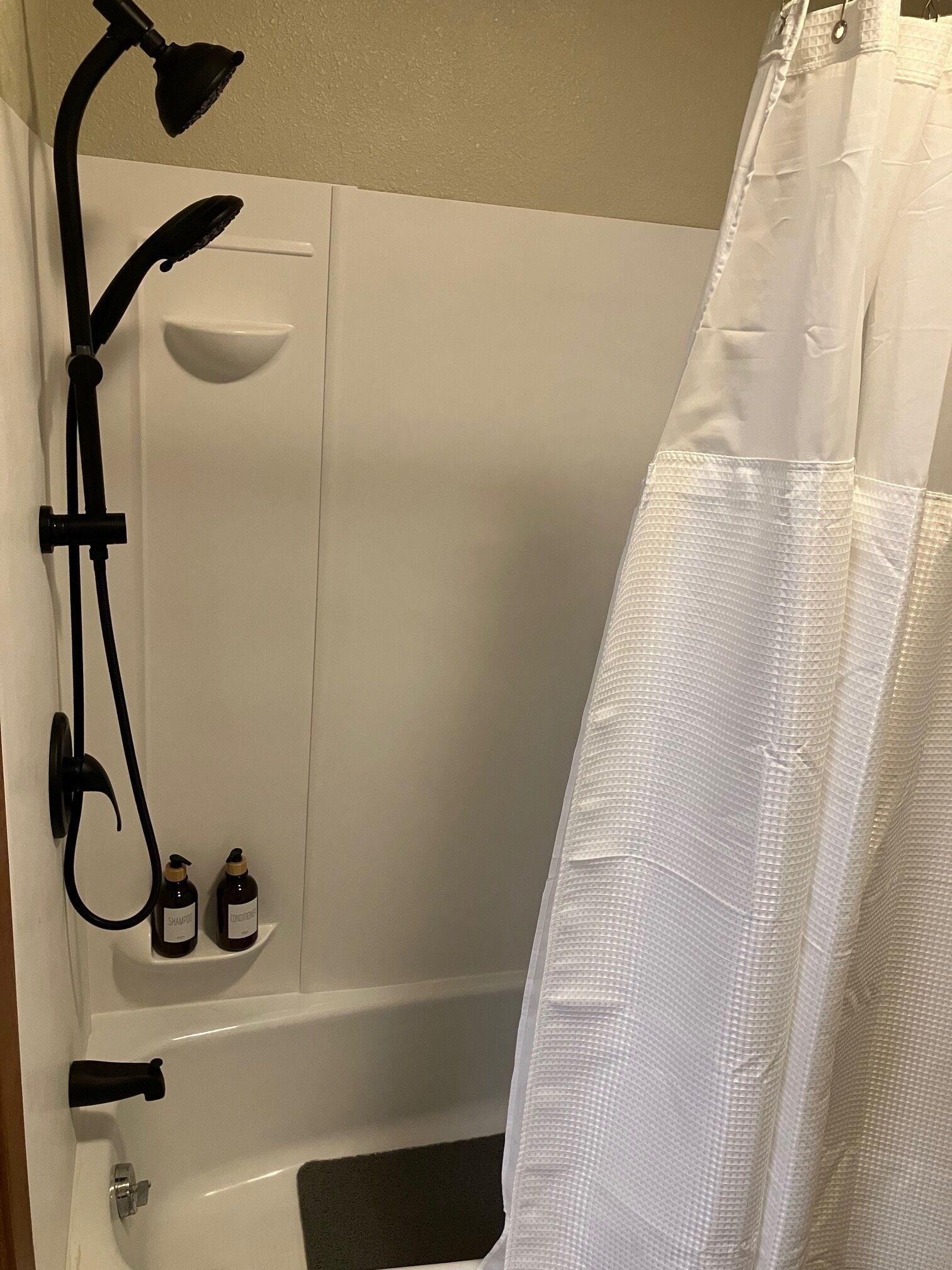 Een badkamer met een witte badkuip, een douche met een handdouche, en twee flessen shampoo en conditioner aan de muur.