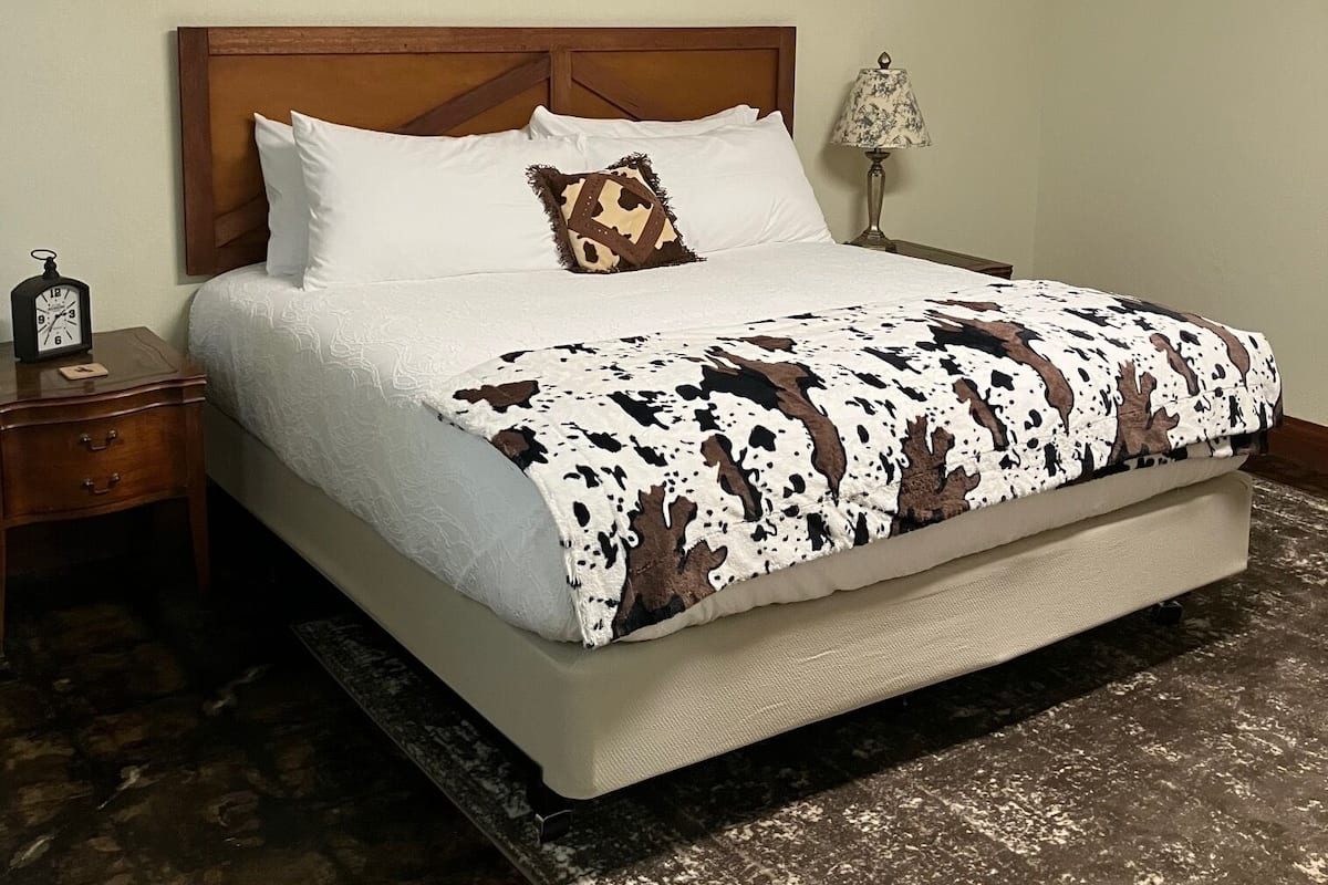 Quarto com cabeceira de madeira, cama com roupa de cama branca e manta estampada em tons de marrom e branco, mesa de cabeceira com relógio e luminária à direita.
