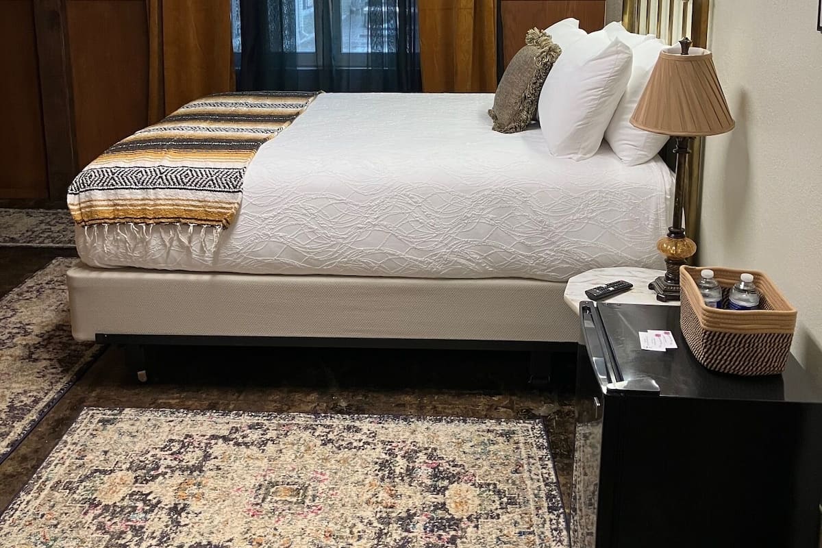 Quarto de hotel bem organizado, com uma cama, mesa de cabeceira, abajur e tapete.
