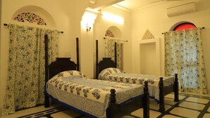 A room with two beds, each with a wooden frame and floral-patterned bedding. There are arched windows with curtains, a ceiling fan, and an air conditioning unit. - Chanod Haveli (Jodhpur)