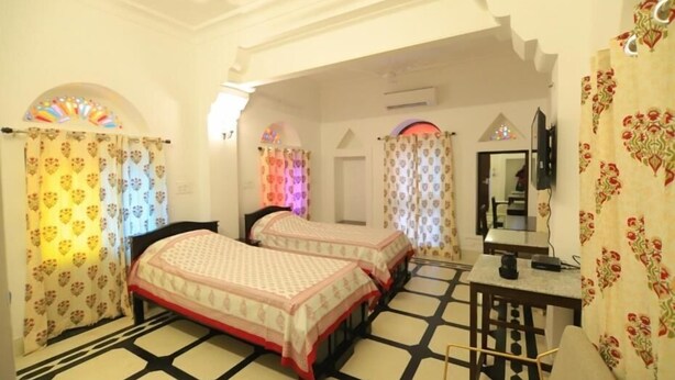 A hotel room with two beds, a small table, a mirror, and a patterned curtain. - Chanod Haveli (Jodhpur)
