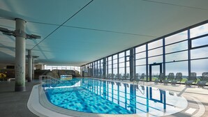 Una piscina interior con agua azul cristalina, rodeada de grandes ventanales que ofrecen vistas del horizonte de la ciudad.