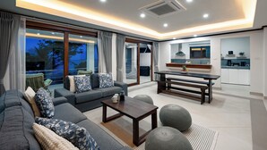 Una sala de estar moderna con un sofá, una mesa de centro y otomanas, una zona de cocina con una mesa de comedor, y grandes puertas correderas de vidrio que dan a un balcón.