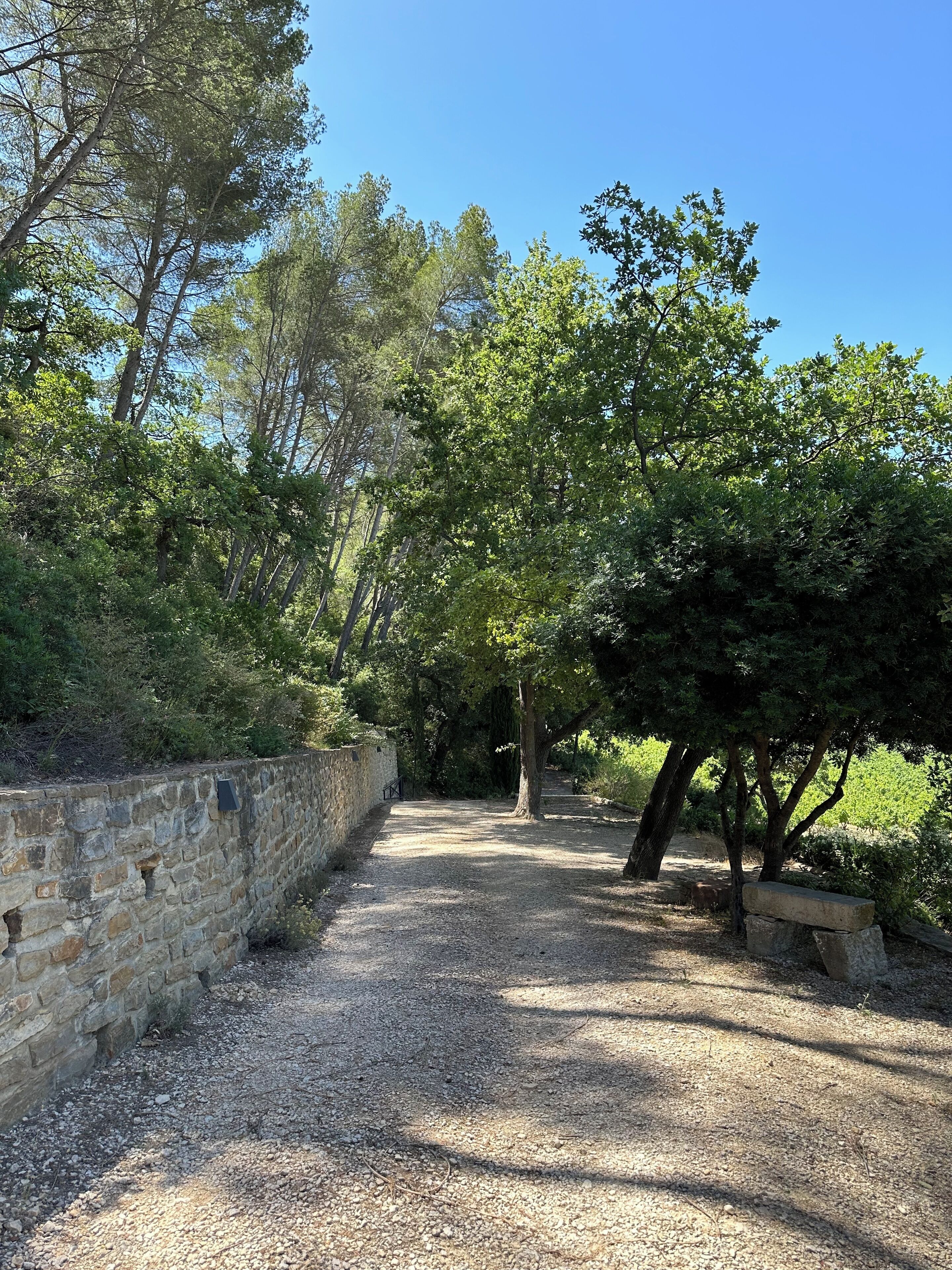 Un sendero de grava bordeado por muros de piedra y árboles, que se adentra en una zona boscosa.