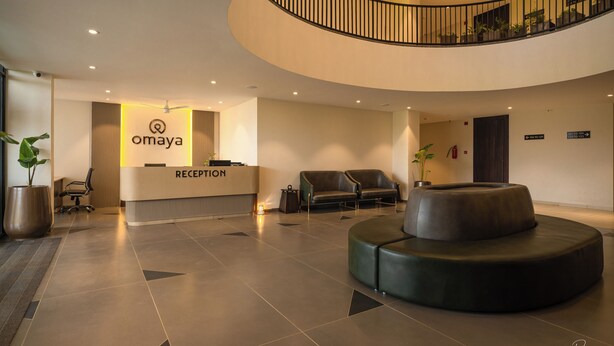 A modern reception area with a reception desk labeled "Omaya," a curved staircase, and a seating area with a round black ottoman. - Hotel Omaya Garden (Raipur)