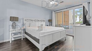 A bedroom with a white bed, a nightstand, a lamp, and a view of the outside. - Legacy Tower II 107 | Charming Ground-Floor Deluxe Unit! (Gulfport)