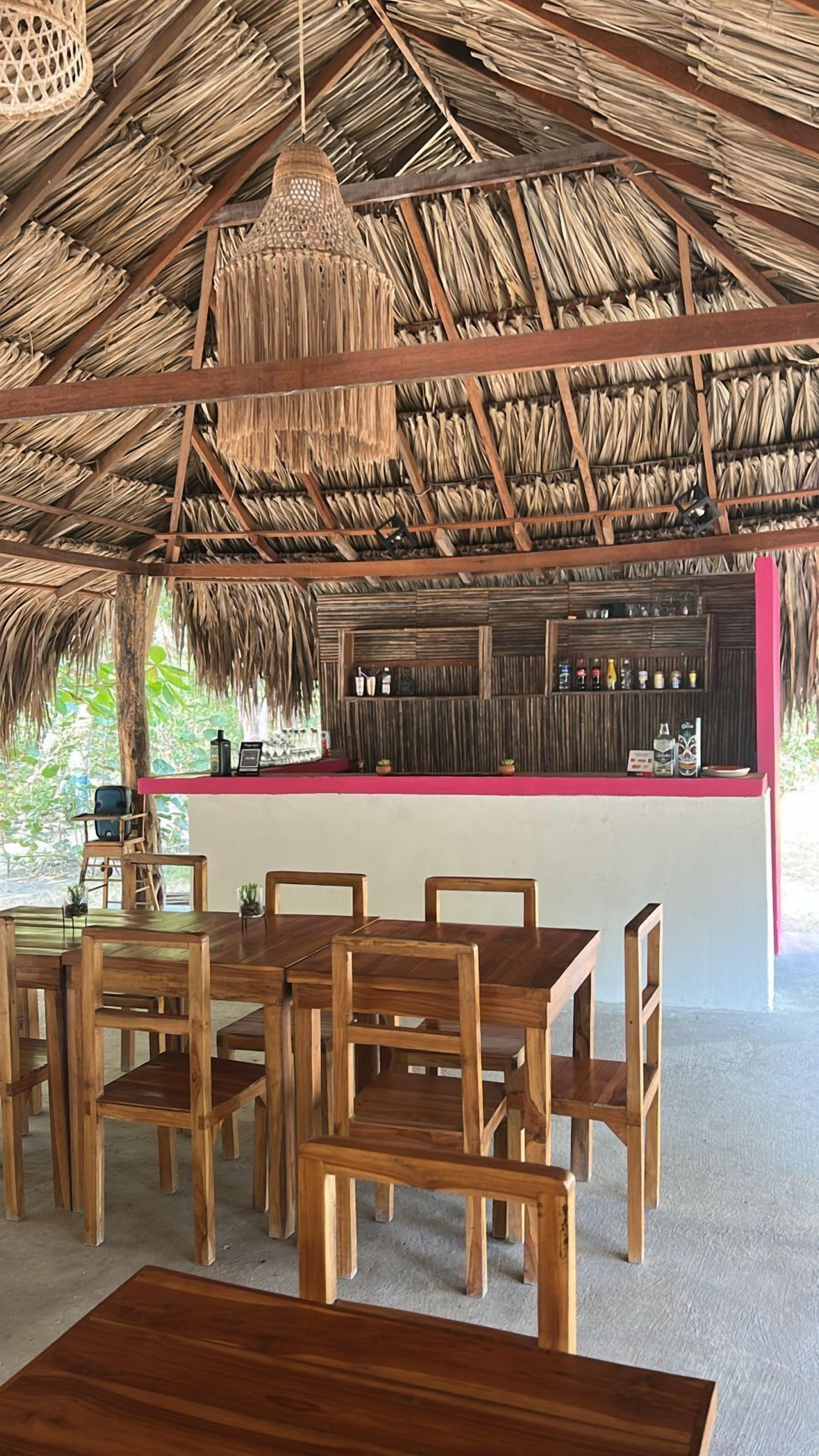 A thatched-roof outdoor dining area with wooden tables and chairs, a bar counter with bottles, and a view of greenery.