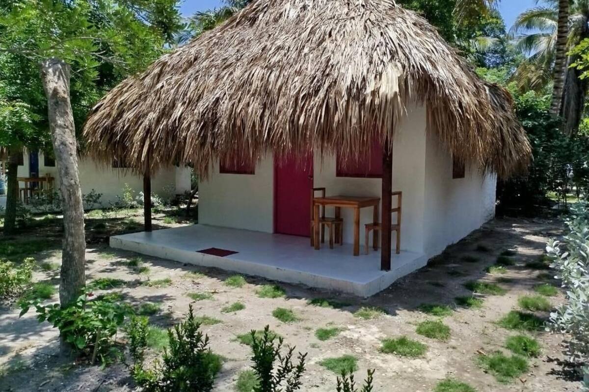 Une hutte au toit de chaume, avec une porte rouge et une petite table et une chaise placées à l’extérieur.