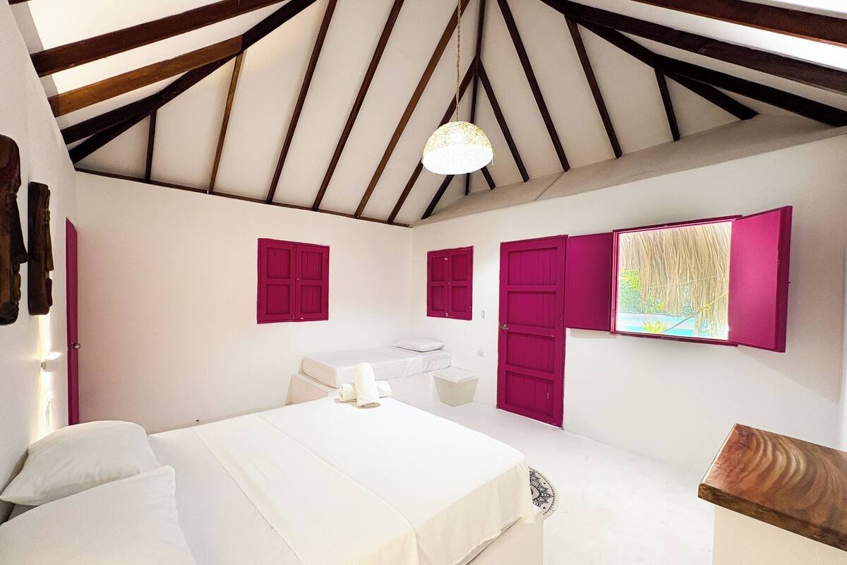 A bedroom with a bed, a small bedside table, a window with a view, and a ceiling with wooden beams.