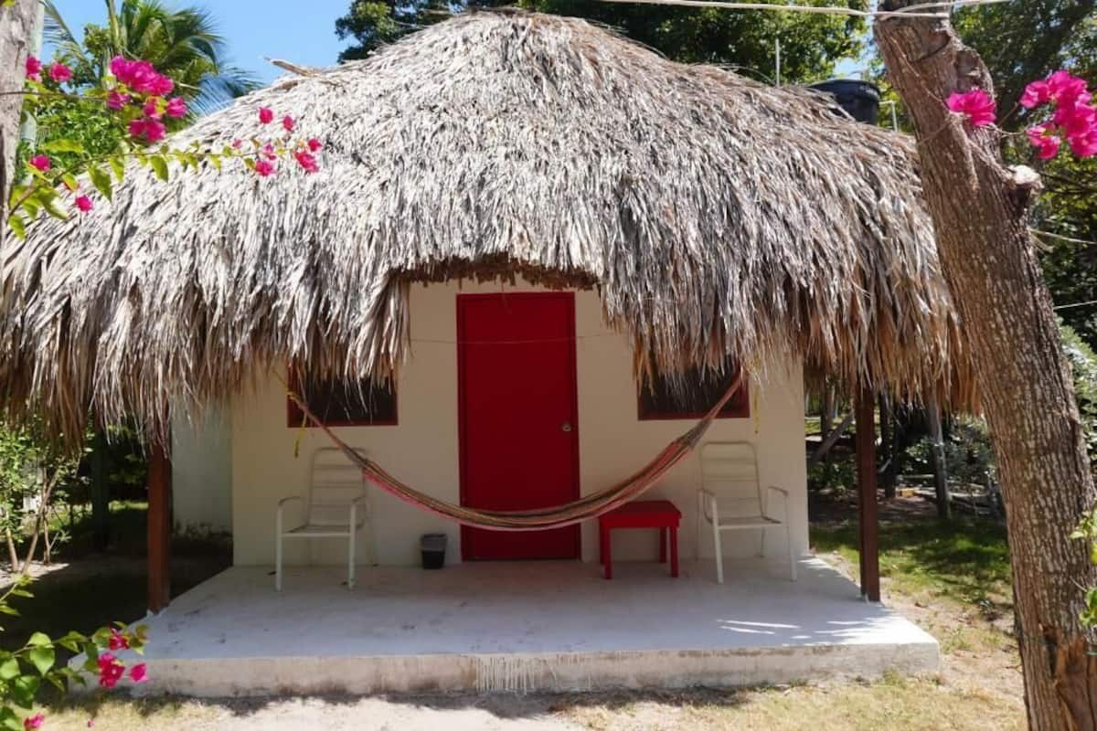 A thatched-roof hut with a red door, two white chairs, and a hammock.