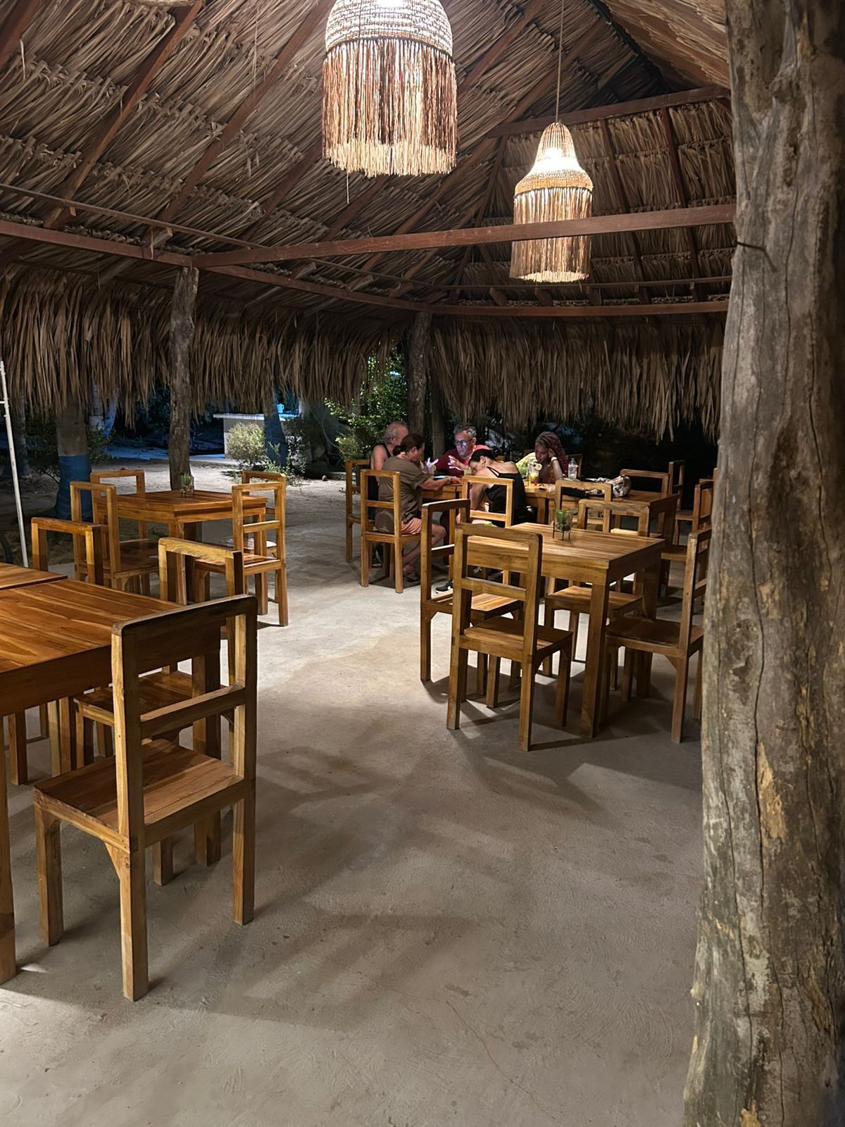 A rustic dining area with wooden tables and chairs, a thatched roof, and hanging light fixtures.