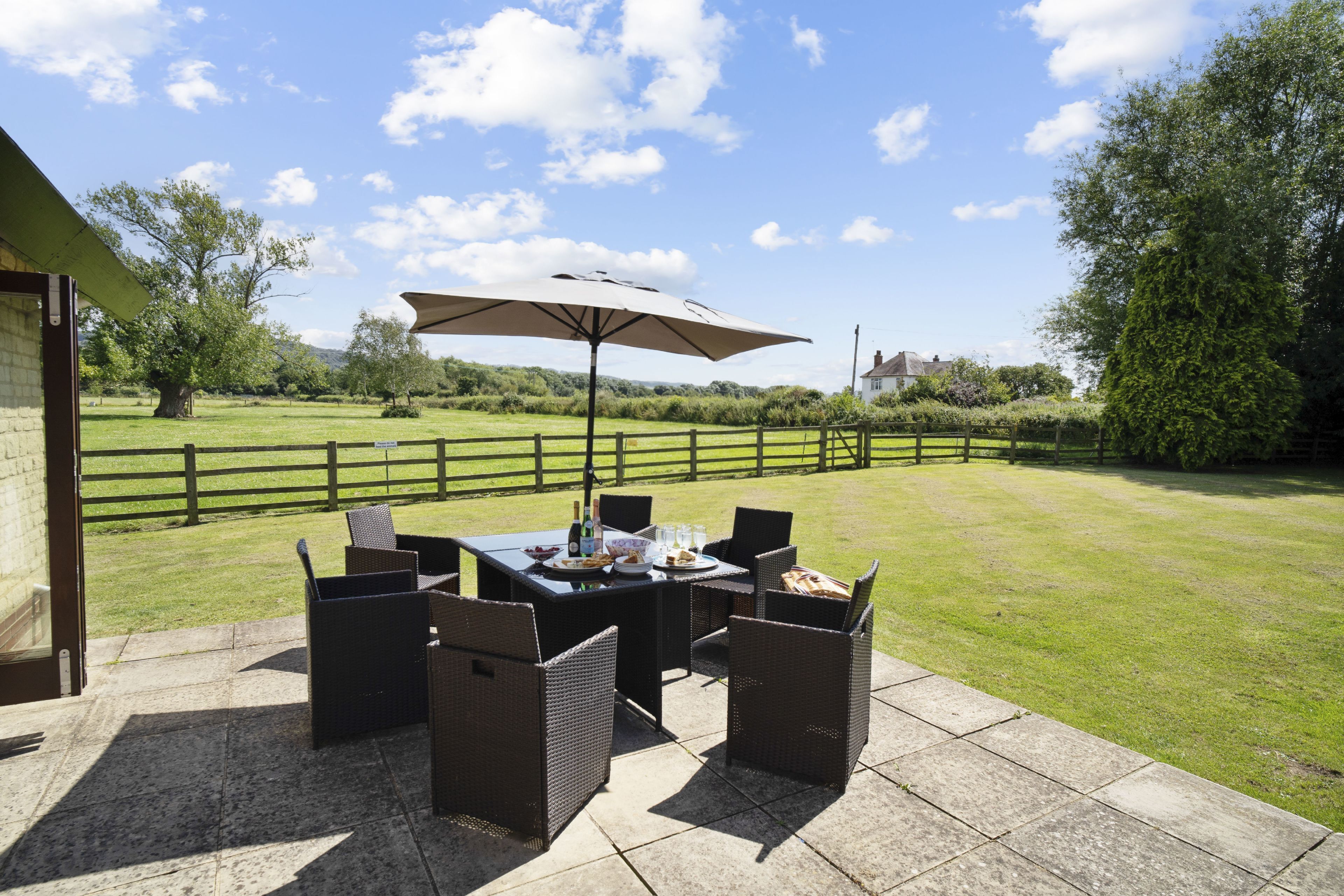 An outdoor patio with a table and chairs, a large umbrella, and a view of a grassy area with trees and a fence.