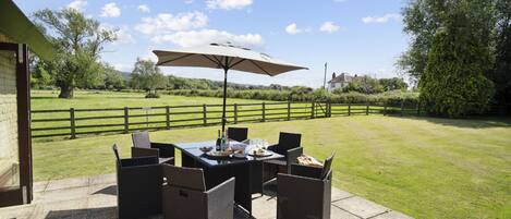 An outdoor patio with a table and chairs, a large umbrella, and a view of a grassy area with trees and a fence.