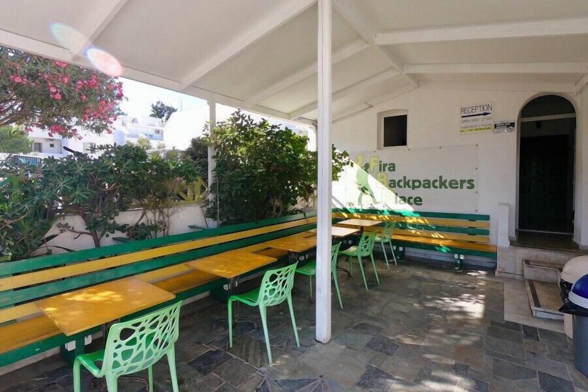 Un espacio al aire libre con bancos y sillas verdes y amarillas, un letrero que dice 'Fira Backpackers Place', y una recepción con una puerta.