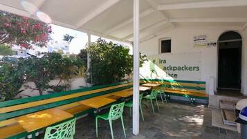 An outdoor seating area with green and yellow benches and chairs, a sign for 'Fira Backpackers Place', and a reception area with a door.