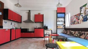 A compact kitchen with red cabinets, a central island, and a dining area with a yellow table and blue sofa. - Fira Backpackers Place (Santorini)