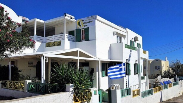 A white building with a balcony, a sign reading "Fira Backpackers Place," and a Greek flag. - Fira Backpackers Place (Santorini)