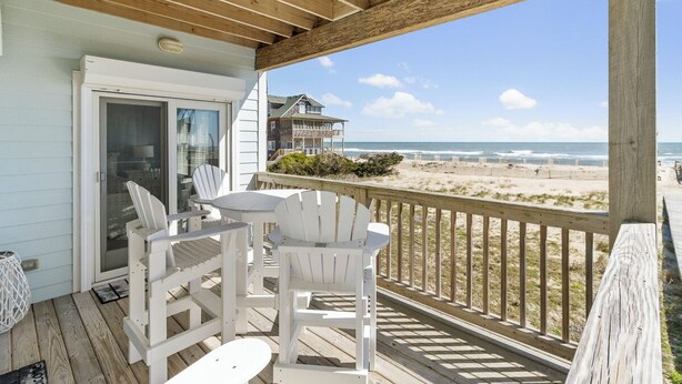 A wooden deck with white chairs and a table, overlooking a beach with a house in the background. - Oceanfront first-floor condo in Rodanthe (Rodanthe)