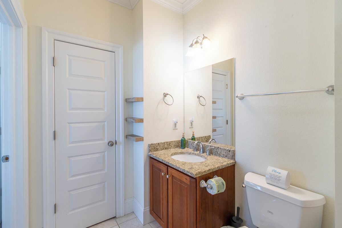 Un baño con puerta blanca, lavabo con encimera de granito, espejo y retrete con dispensador de toallas de papel.
