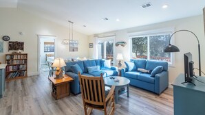 A spacious living room with two blue sofas, a wooden chair, a small round table, and a bookshelf. - Sun Realty pet-friendly oceanside home just 500' to the beach (Duck)