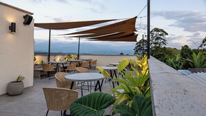 Une terrasse extérieure avec des tables rondes et des chaises en osier, entourée de grands pots de plantes et offrant une vue sur les montagnes lointaines.