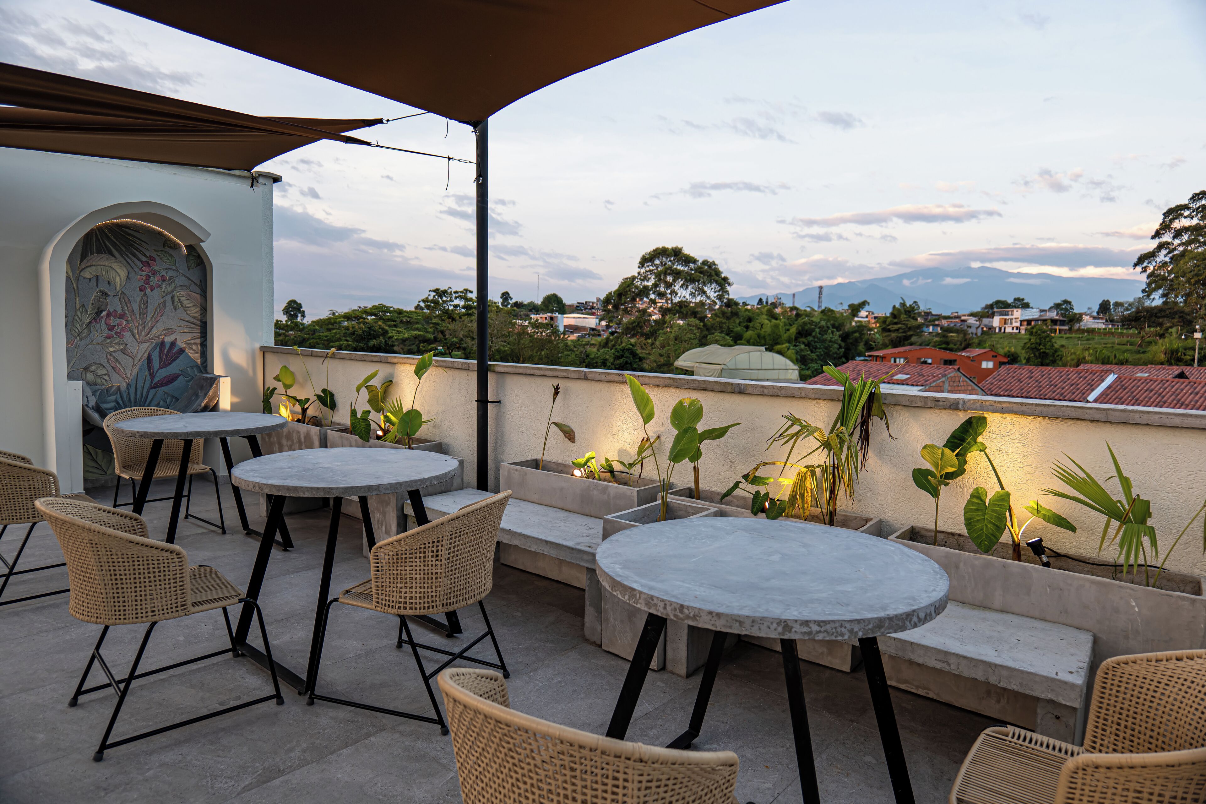 An outdoor patio with round tables and wicker chairs, surrounded by potted plants and a view of the cityscape in the background.