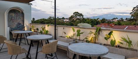 An outdoor patio with round tables and wicker chairs, surrounded by potted plants and a view of the cityscape in the background.