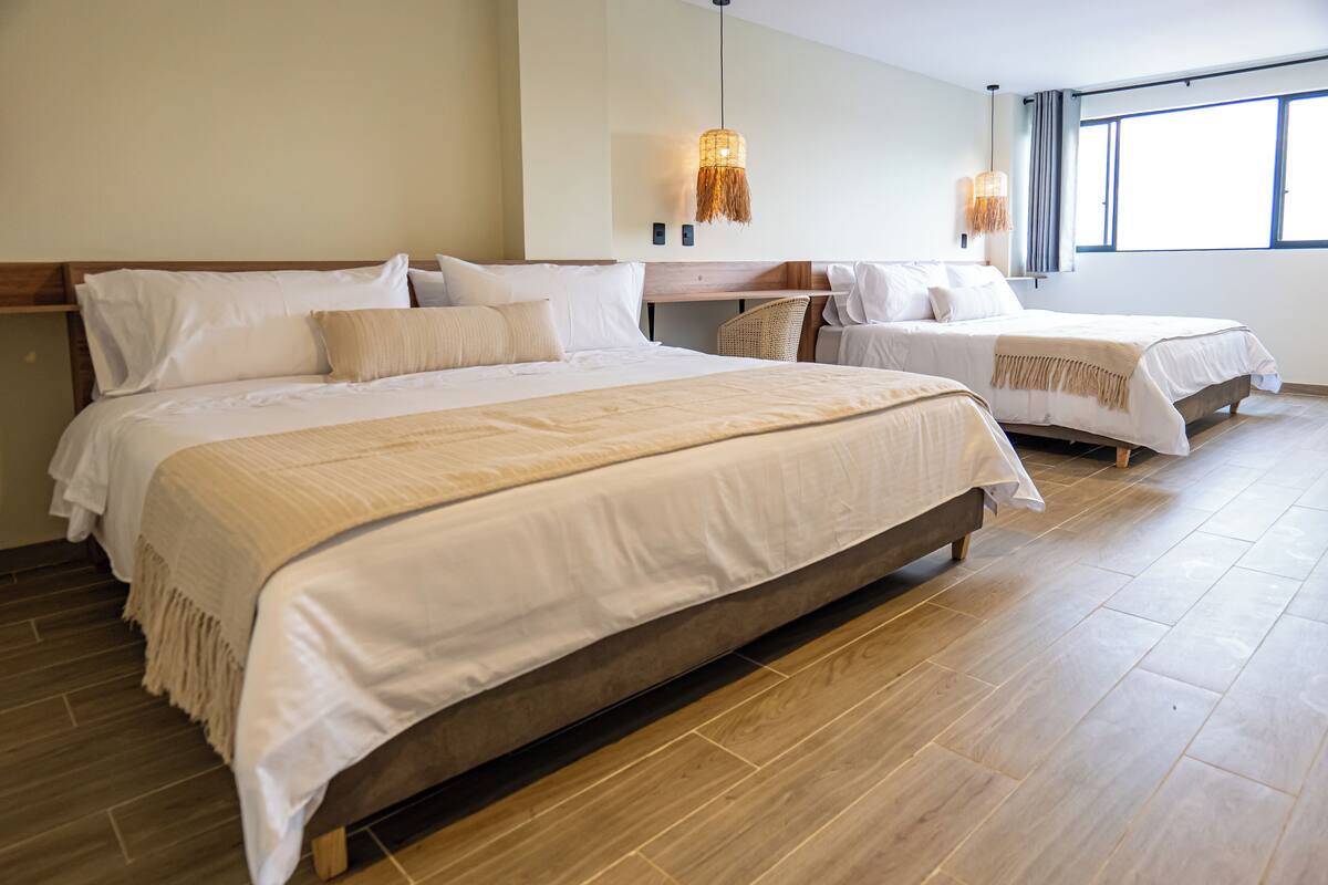 A hotel room with two beds, a wooden floor, and a large window providing natural light.