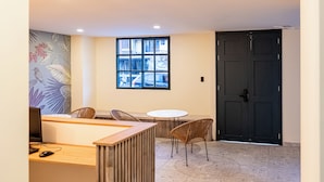 Un espace d’accueil avec un bureau en bois, deux fauteuils en osier, une table ronde, une grande fenêtre donnant sur la rue et une porte sombre.
