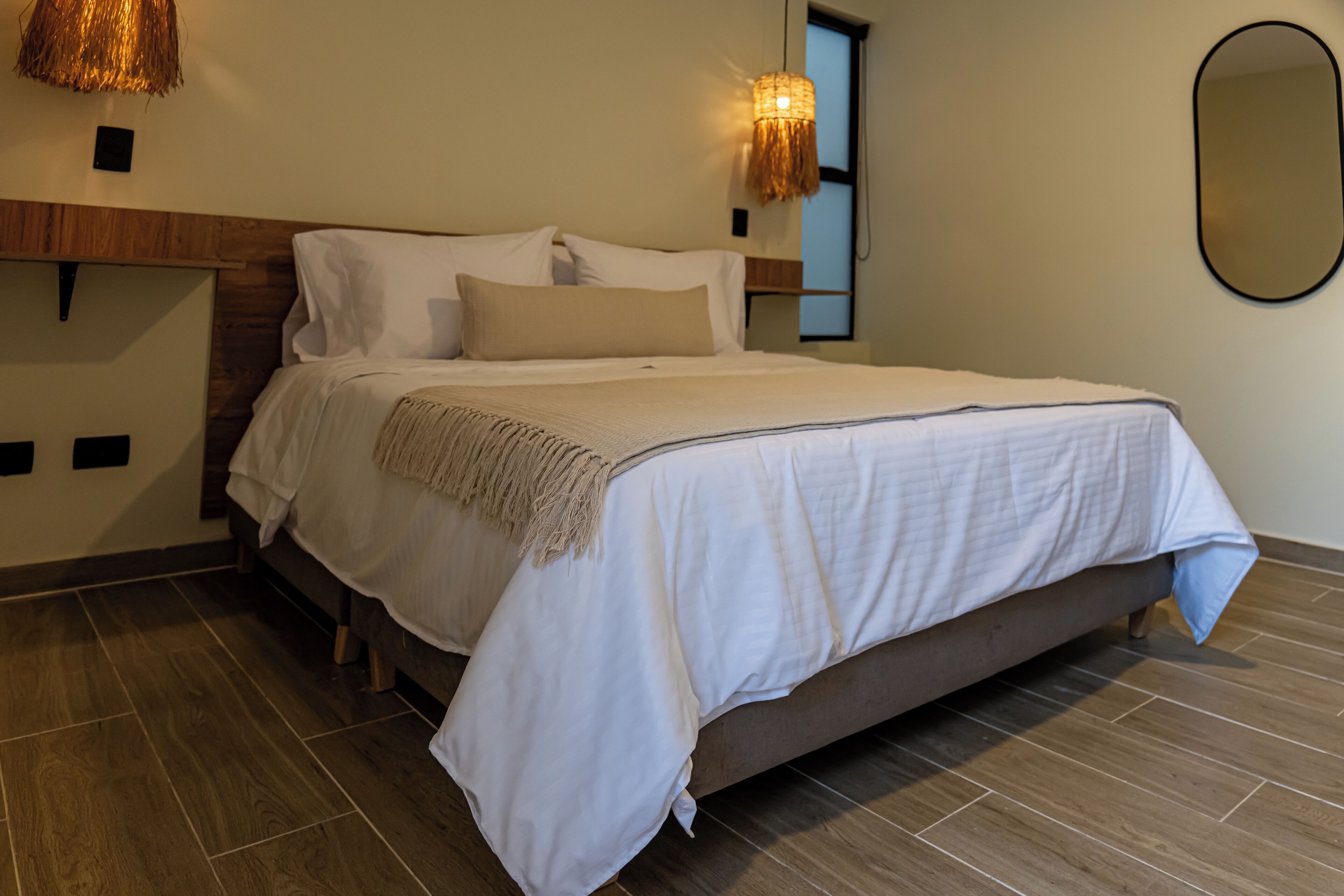 A neatly made bed with a wooden headboard, a woven light fixture above it, and a large mirror on the wall.