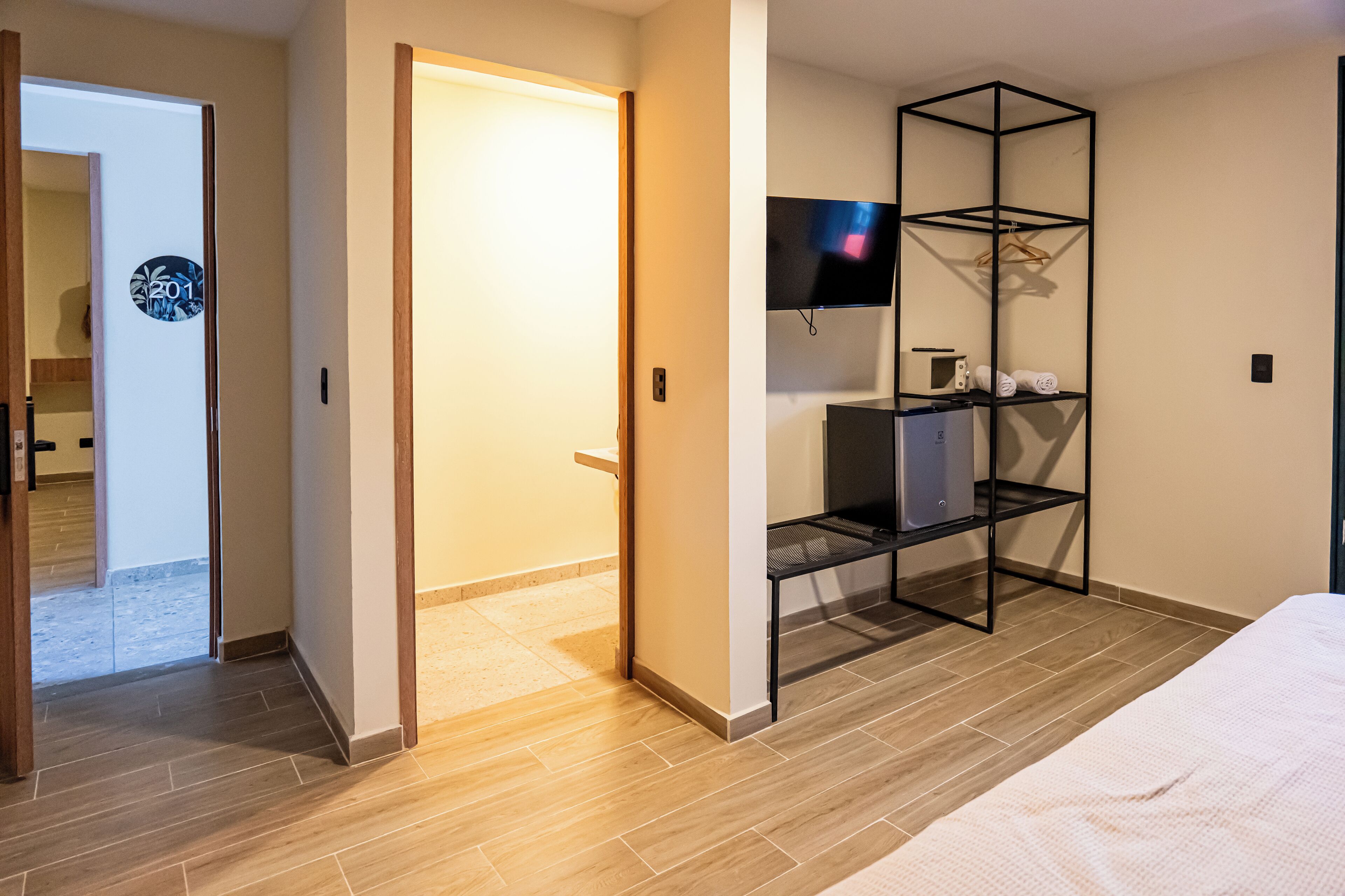 Een hotelkamer met een houten vloer, een bed, een televisie op een zwart metalen plank, en een deur naar een andere kamer.