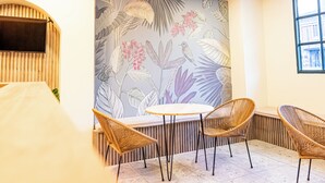 Une pièce avec une table ronde et quatre chaises en osier, une fresque représentant des feuilles et des fleurs tropicales, et une fenêtre donnant sur des immeubles.