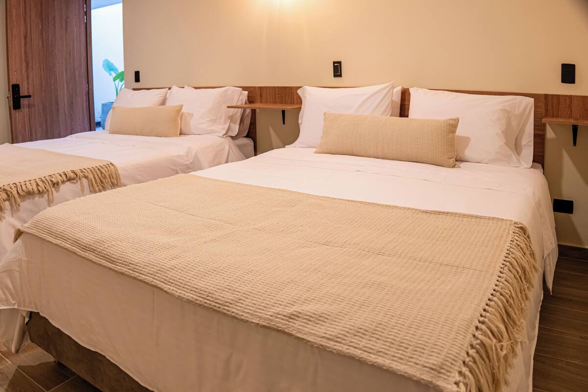 A hotel room with two beds, each with white linens and beige throw blankets.