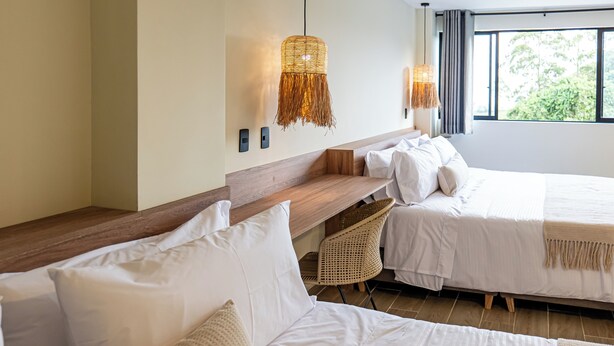 Une chambre d’hôtel avec deux lits, une tête de lit en bois, un luminaire en fibres tressées et une vue sur la verdure.