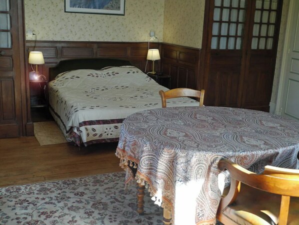 Un dormitorio con una cama, dos lámparas de noche, una silla de madera y una mesa con mantel estampado.