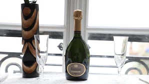 A bottle of Ruinart Champagne, two glasses, and a wooden vase on a table by a window.