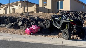 Zona residencial con una casa, un coche de juguete rosa y un vehículo todoterreno estacionados adelante.