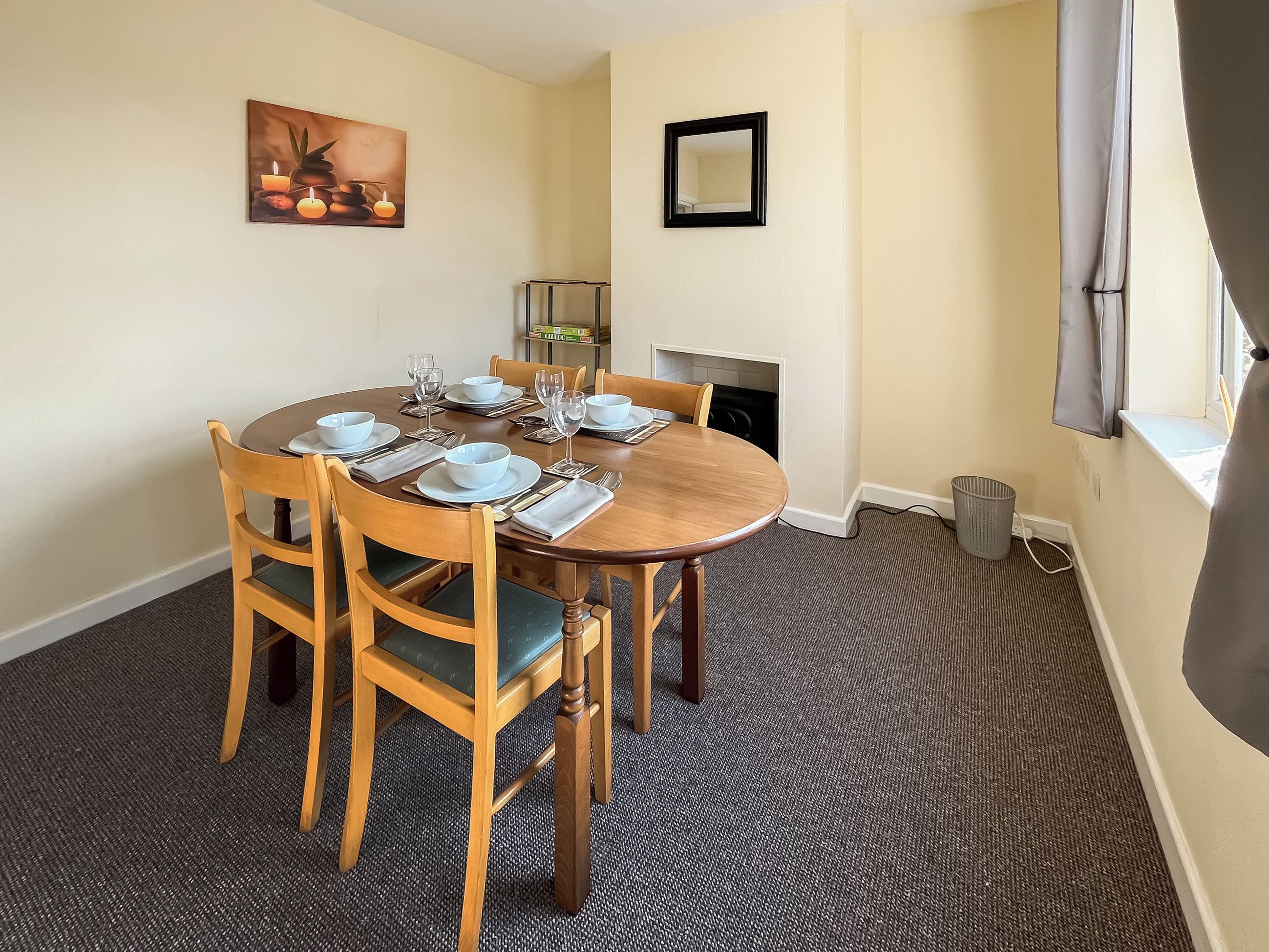A dining room with a round table set for four, wooden chairs, a painting on the wall, a mirror, and a fireplace.