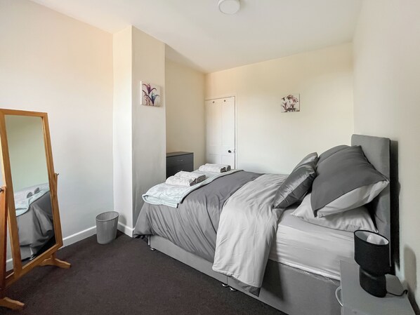 A bedroom with a bed, a bedside table, a mirror, a trash bin, and a wall-mounted coffee maker.