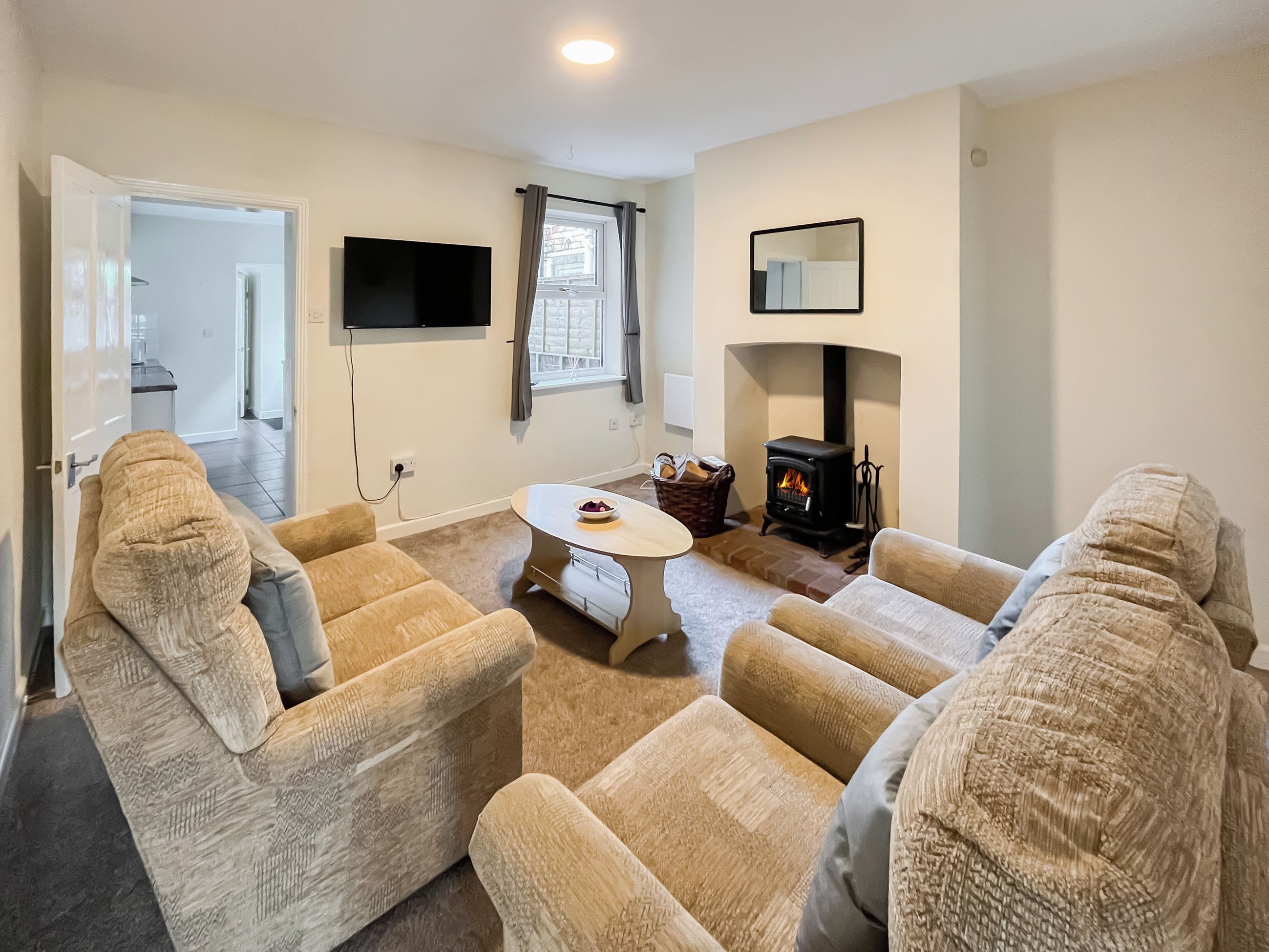 A living room with beige sofas, a round coffee table, a fireplace, a TV mounted on the wall, and a door leading to another room.