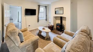 A living room with beige sofas, a round coffee table, a fireplace, a TV mounted on the wall, and a door leading to another room.